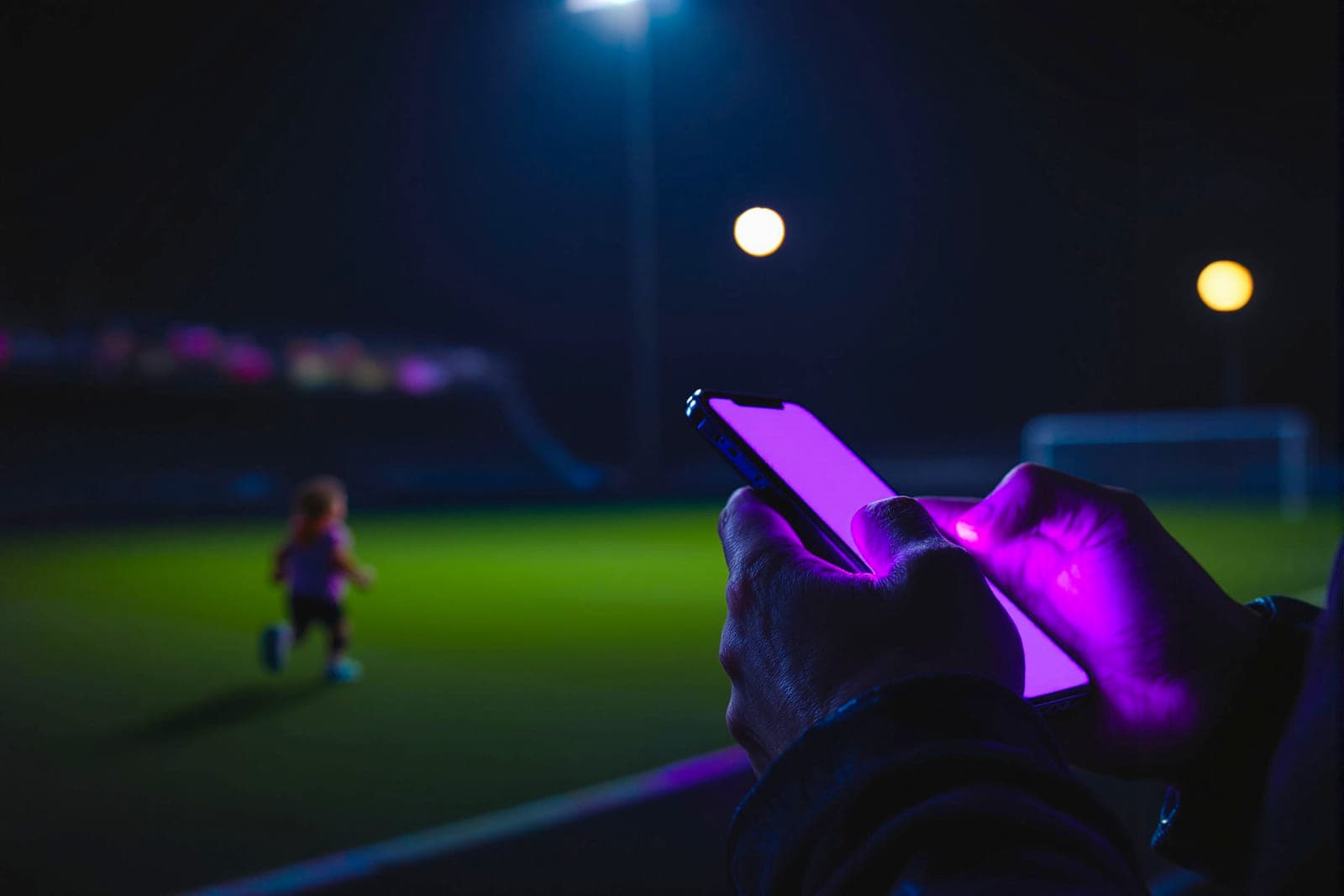 An adult holding a phone at the side of a pitch, the screen the only light.